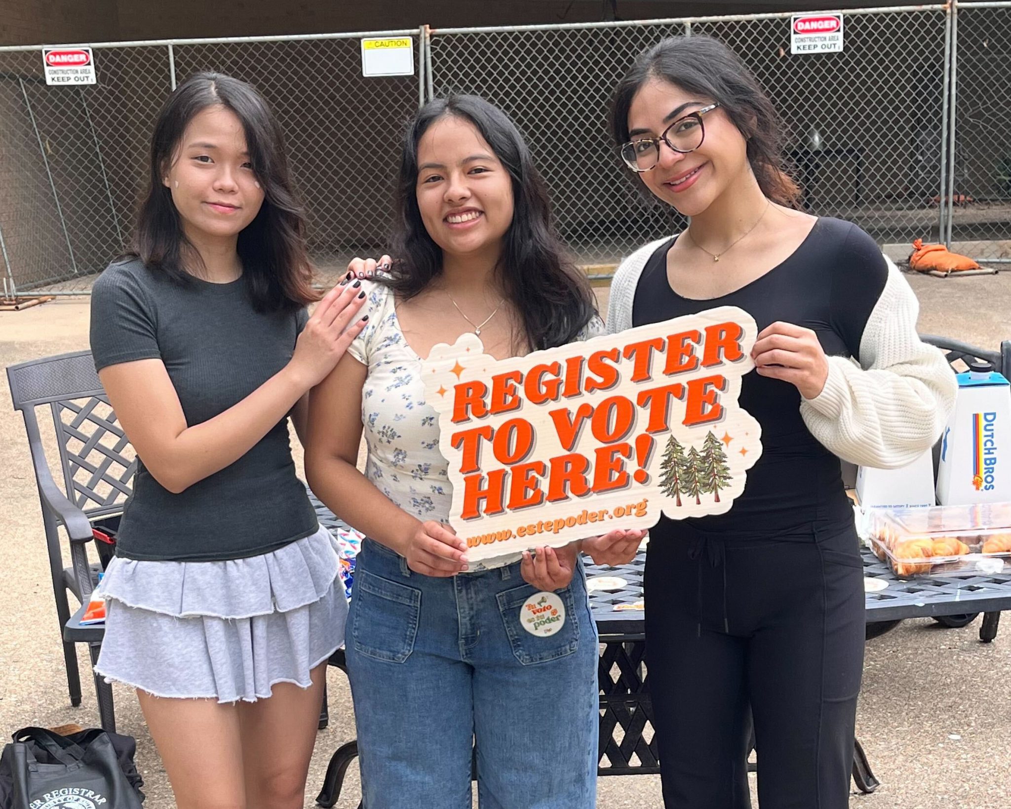 Three women stand together with a sign that reads "Register to Vote Here"